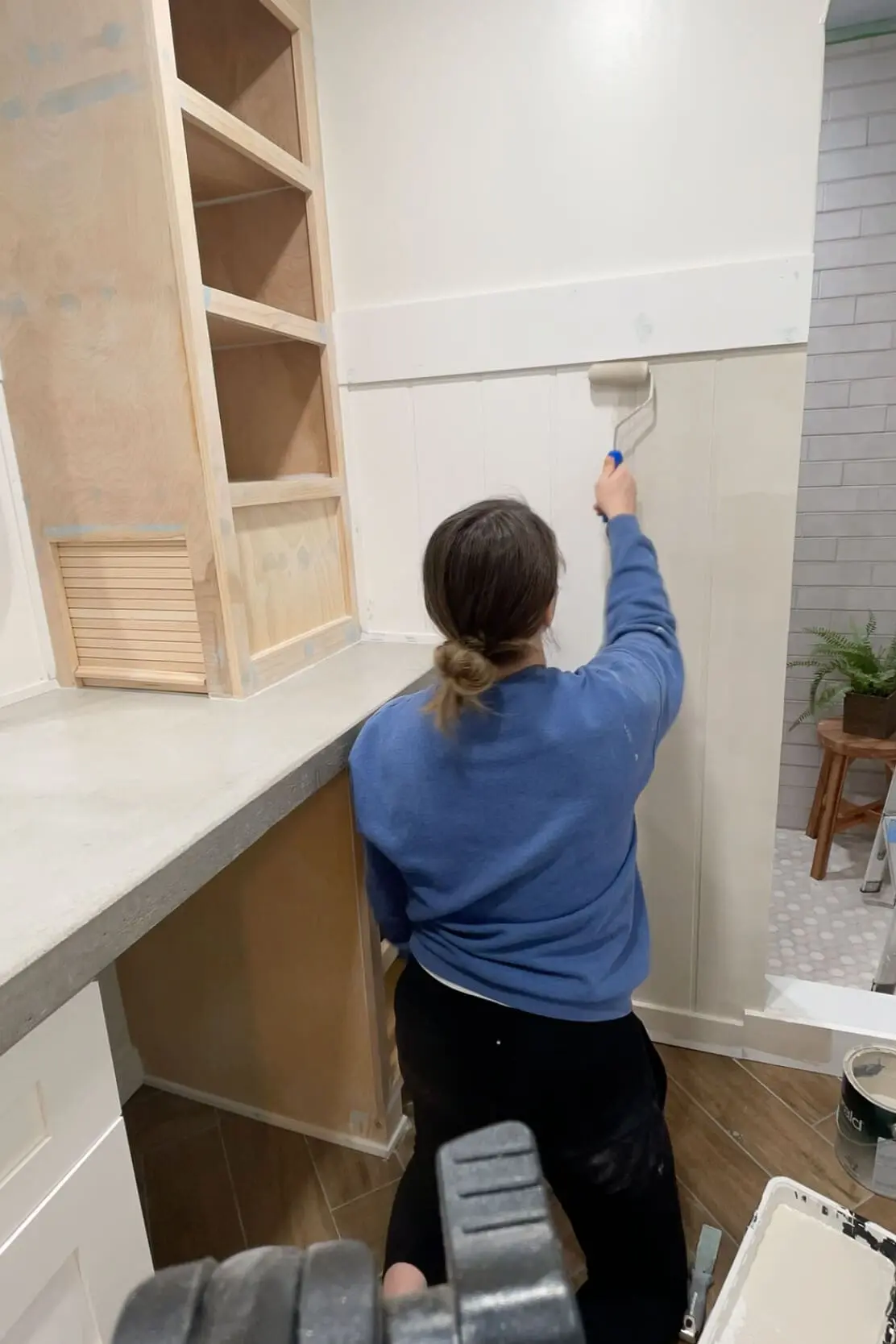 A woman painting a white shiplap accent wall in a bathroom with a roller brush. The space features a custom-built wooden cabinet, a concrete countertop, and an arched shower entrance, showcasing the transformation in progress.